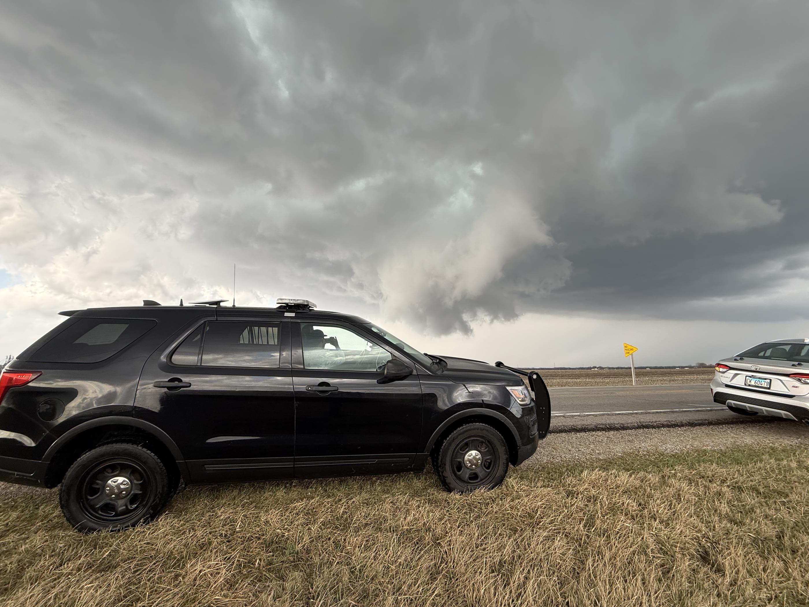 The TwistedWX chase vehicle parked on a grass shoulder with a lowering storm base and rain shaft behind it.