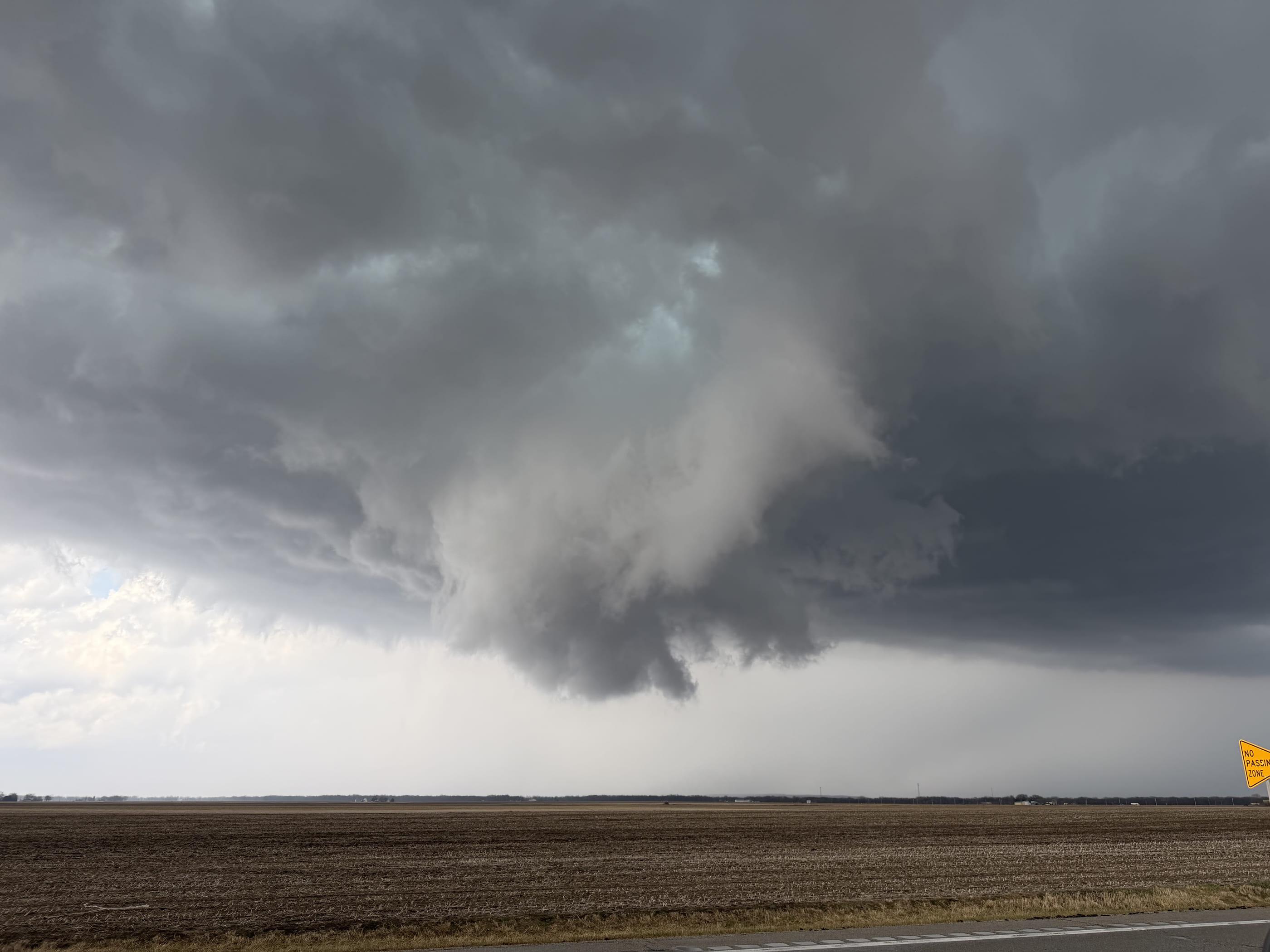 A broad, ragged wall cloud hanging beneath a dark storm base over bare farm ground, photographed from a roadside in north-central Illinois.