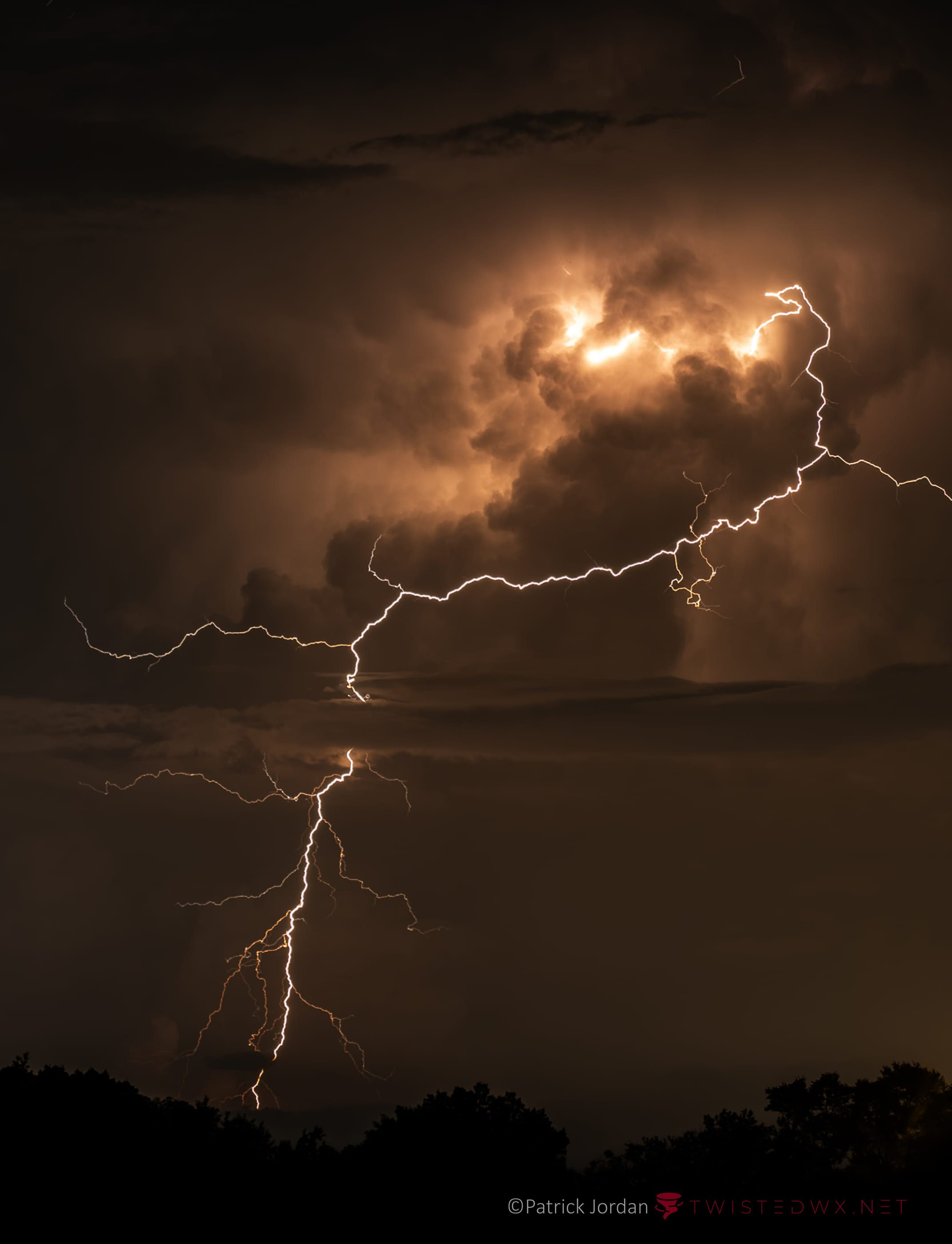 Branching lightning across a thunderstorm lit orange from within, above a dark treeline.