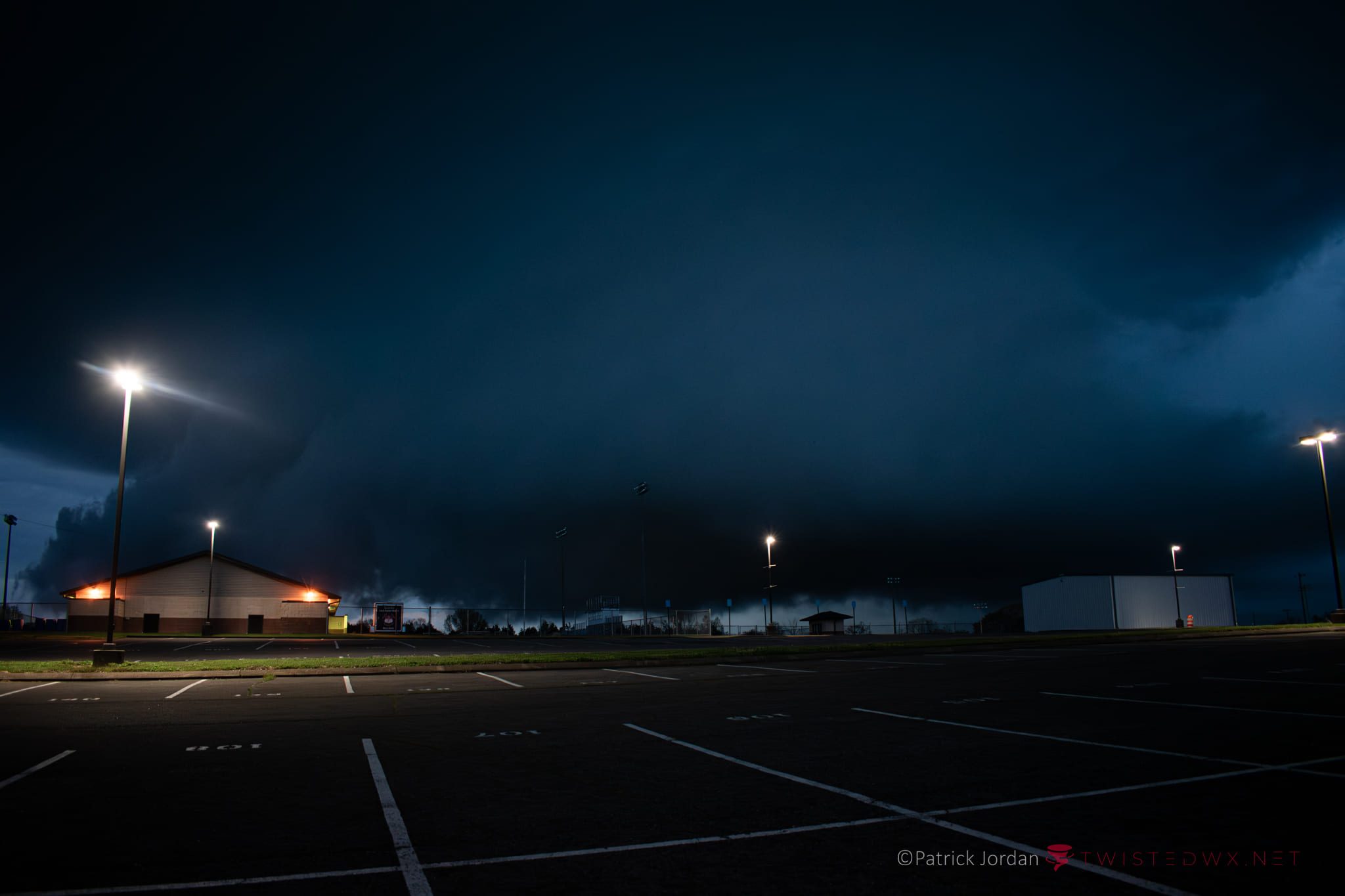 Low storm structure moving over an empty, floodlit parking lot after dark.